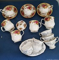 Top view of Royal Albert Old Country Roses and Silver Birch cups, saucers, creamer, sugar bowl, candlestick lid, and serving plate arranged on dark blue fabric background.