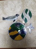Photo shows four stained glass decorative pieces including a bird, leaf, round abstract panel, and star shown on a fabric background with eyeglasses for scale.