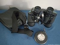 Photo showing black Tasco binoculars with textured grips and adjustable shoulder strap, and a vintage Hoppe tape measure with black composite case.