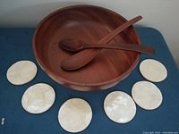 Photo showing one large wooden salad bowl with two wooden utensils inside and six natural Capiz shell coasters arranged around the bowl.