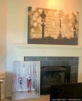 Two large canvas art prints above and in front of fireplace, showing the Eiffel Tower print on the wall and the smaller French scene canvas in front leaning on the fireplace.