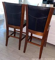 Back view of two faux leather high chairs showing black upholstery and brown wood legs and trim.