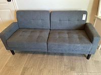 Front view of gray upholstered couch with tufted seat and back cushions, wooden legs, and slight wear.