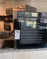 Front view showing black Magic Mill food dehydrator with removable racks inside and Taylor digital food scale on countertop.