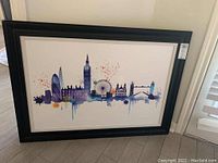 Front angled view of black framed London skyline watercolor art with visible landmarks and paint splashes.