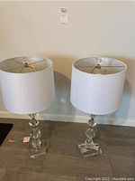 Two acrylic table lamps with white drum shades on wood floor against wall.