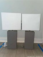 Front view of two rectangular table lamps with grey faux leather bases and white rectangular fabric shades placed side by side on floor.