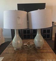 Pair of teardrop-shaped mirrored acrylic base lamps with white linen drum shades placed on a wooden table in front of a TV stand.
