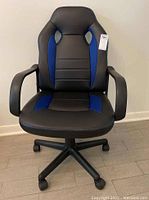 Front view of black office chair with blue trim, armrests, and caster wheels on a wood floor against a white wall.
