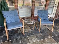 Two wicker rocking chairs with blue cushions and matching round wicker side table with glass top on tiled floor in front of windows.