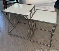 Photo showing two square side tables with metal frames and white faux marble-look tops, one larger rectangular and one smaller square, placed on wooden floor near couch.