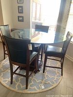 Photo showing round glass top dining table surrounded by four high wood and leatherlike chairs, all on a round patterned rug near windows.
