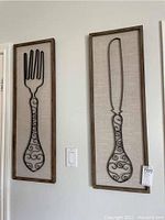 Two framed metal wall art pieces shaped like kitchen utensils hung on a wall.