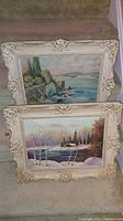 Two framed landscape paintings stacked showing coastal and winter scenes with ornate frames.