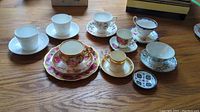 Wide shot of the entire collection showing 7 teacups, 7 saucers, 1 plate, and a small decorative dish on hardwood floor.