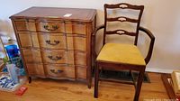 Four drawer wooden dresser with curved front and metal handles next to ladder back wooden chair with yellow upholstered seat