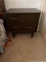 Front view of one dark wood night table showing two drawers with large curved handles and tapered legs, positioned on carpeted floor.