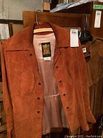 Front view of rust-colored suede and leather jacket with snap buttons, hanging on a wooden hanger and chair