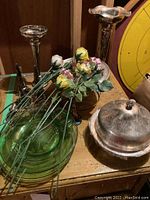 Two tall silver plated candle holders, silver plated chafing bowl with lid, green glass plates, and ceramic flowers on wooden surface