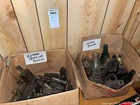 Two open cardboard boxes filled with assorted vintage glass bottles, some green, brown, and clear. Boxes show wear. Bottles are various sizes and shapes with some labeling visible.