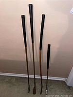 Four vintage golf clubs standing against a wall, showing a variety of shaft types and club heads