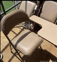 Side view of a taupe steel folding chair with padded seat, backrest and tablet arm extended