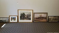 Five framed artworks arranged on floor against wall showing variety of themes including horse racing scenes, floral imagery, and landscapes. Frames range in size and style.