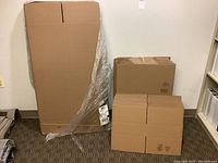 Three different sizes of unused flattened cardboard boxes, stacked and leaning against a wall, including a large long narrow box on the left and smaller boxes stacked on the right.