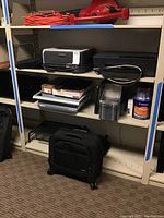 Shelf with Brother HL-2140 printer, HP Officejet Pro 6230 printer, Fellowes Quasar 500 binding machine, blank DVDs, packing material, and Samsonite carry on bag below.