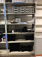 Overall shelving with multiple plastic storage bins, office supplies, and filing accessories including calculators, staplers, and papers.
