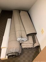 Rolled commercial carpet remnants beige color in various sizes with wood molding pieces on brown carpet floor.