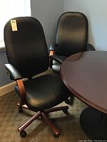 Two matching black leather office chairs with wood armrests positioned near a round wooden table. Chairs show wear on wood components.