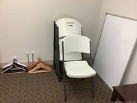 Six folding chairs stacked against the wall, four white chairs visible, whiteboard leaning nearby, hangers on floor