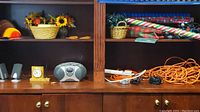 Shelf with Thanksgiving floral basket, Christmas string lights, gift wrap rolls, two computer speakers, small yellow desk clock, and orange extension cords