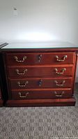 Front view of the wooden file cabinet showing four drawers, brass handles, lock on two drawers, and glass top.