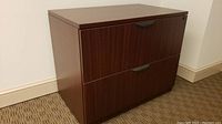 Front angle photo of two-drawer laminate file cabinet with brown wood-grain finish placed against a light wall on carpeted floor.