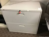 Front view of white metal two-drawer file cabinet with lock and keys on top drawer, showing scratches and wear