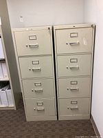 Two putty colored Hon brand metal file cabinets side by side, each with four drawers, label holders, and handles. Visible minor scratches and paint marks on cabinet surfaces.