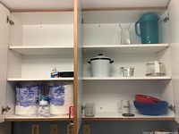 Kitchen cabinet with paper plates, napkins, plastic bowls, glass measuring cup, coffee carafe, and plastic pitcher.