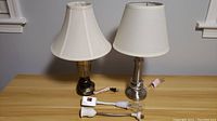 Photo of two metal table lamps with beige fabric lampshades and two plug-in bulb sockets on a wooden surface.