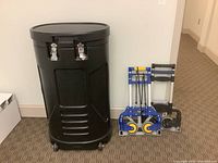 Front view of large black oval carrying case on wheels beside two luggage carts: one blue with yellow wheels and one black marked Magna Cart