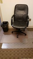 Black adjustable rolling office chair on clear vinyl carpet mats with black plastic waste bin next to it
