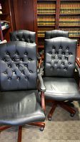 Photo showing front view of four black leather office chairs with button tufting and wooden armrests with silver studs. Chairs have caster wheels and are arranged in a room with bookshelves.