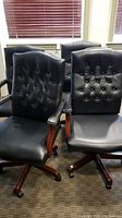 Four matching black office chairs with button tufted backs, wooden studded arms and caster rolling bases in an office room environment.
