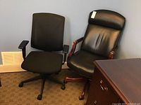 Two office chairs side by side: one black fabric mesh chair with adjustable arms and one black leather chair with wood armrests and base, showing some wear.