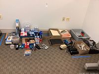 Wide photo showing spread of office supplies on floor including boxes, speakers, folders, wires, staplers, tape dispensers and other small desk items.