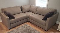 Overall view of beige L-shaped sectional sofa with dark gray throw pillows in living room near window and hardwood floor