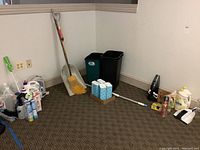 Wide view showing trash cans, broom, shovel, vacuum, and various cleaning products arranged against the wall on carpeted floor.