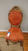 Front view of the vintage style chair showing ornate gold curved wooden frame with crown floral detail at top, coral upholstery with leaf pattern, and front legs with small caster wheels.
