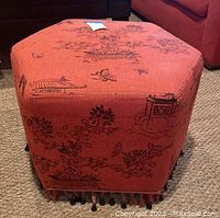 Front view of reddish orange hexagonal ottoman with black chinoiserie floral and architectural design, tassel beads at bottom edge.